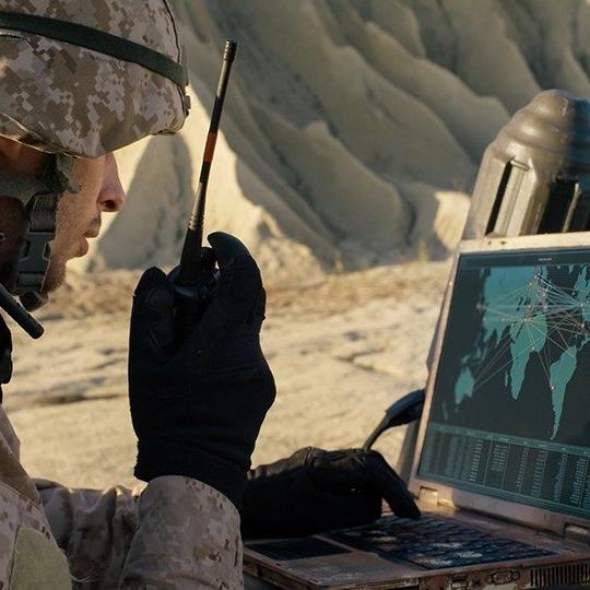 Man dressed in combat gear talking on a radio while looking at a map of the world on a miltary laptop