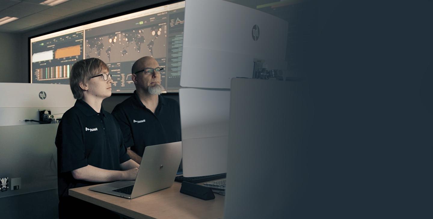Two male Viasat employees wearing black polos featuring a white Viasat logo working in a cybersecurity operations center